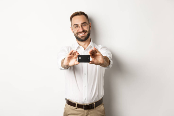 Handsome man in glasses holding a credit card, smiling pleased, standing over white background - Fotó, kép