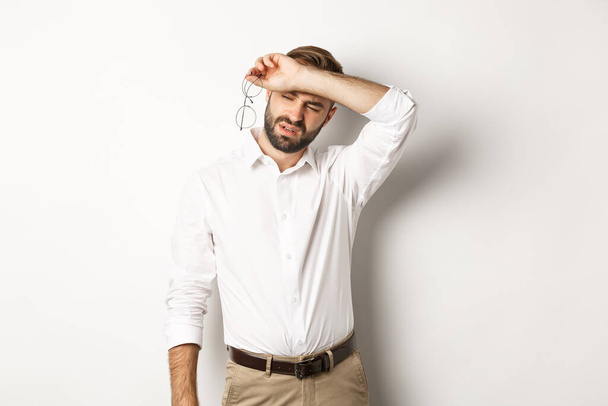 Уставший офисный работник снимает очки, вытирая пот с лба рукой, стоя осушенный на белом фоне - Фото, изображение