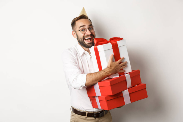 Feiertage und Feiern. Aufgeregter Mann, der Geburtstagsparty feiert und Geschenke erhält, glücklich aussehend, vor weißem Hintergrund stehend - Foto, Bild
