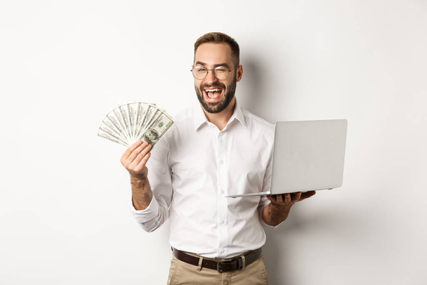 Бизнес и электронная коммерция. Уверенный бизнесмен показывает, как работают в Интернете, подмигивая, держа деньги и ноутбук, стоя на белом фоне - Фото, изображение
