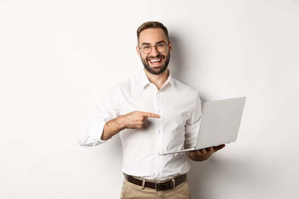 Obchod. Hezký manažer v brýlích pracuje na notebooku, ukazuje na počítač a usmívá se potěšen, stojí nad bílým pozadím - Fotografie, Obrázek