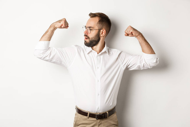Exitoso hombre de negocios flex bíceps, mostrando los músculos y buscando confiado, sintiéndose fuerte, de pie sobre fondo blanco - Foto, imagen