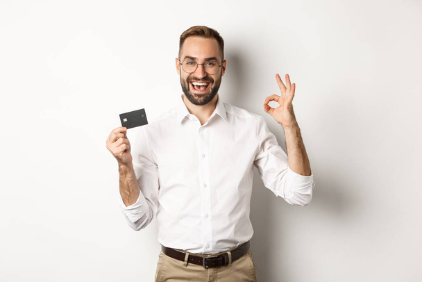 Un bell'uomo che mostra la sua carta di credito e il cartello ok, raccomanda banca, in piedi su sfondo bianco - Foto, immagini