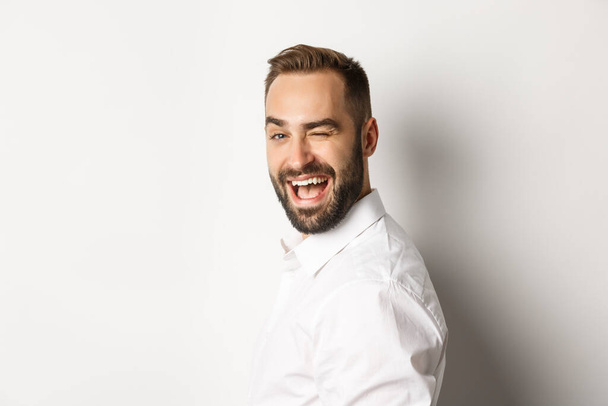 Close-up of cheeky guy with beard, turn face at camera and winking with smile, standing over white background - Foto, immagini