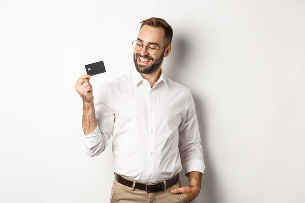Knappe zakenman met zijn credit card, op zoek tevreden, staande over witte achtergrond - Foto, afbeelding