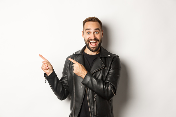 Homem macho confiante em jaqueta de couro preto, apontando os dedos deixados na oferta promo, mostrando logotipo, fundo branco - Foto, Imagem