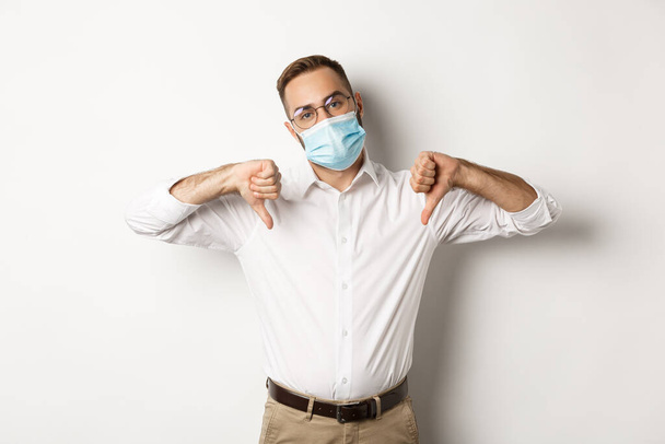 Covid-19, social distancing and quarantine concept. Displeased boss wearing face mask and showing thumbs down, dislike and disapproval sign, white background - Fotoğraf, Görsel