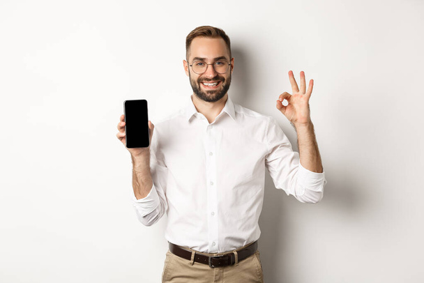 Gestor joven satisfecho mostrando la pantalla del teléfono inteligente y el signo de aprobación, recomendando la aplicación, de pie sobre fondo blanco - Foto, Imagen