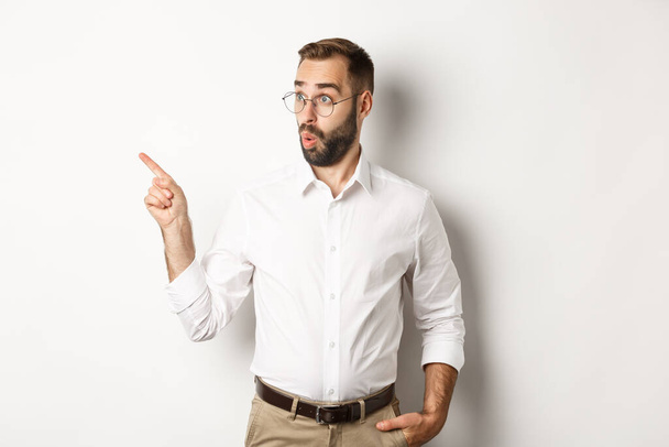 Zaintrygowany młody człowiek w okularach wskazujących palcem w lewo, patrząc na ofertę promocyjną zainteresowany, stojąc na białym tle - Zdjęcie, obraz