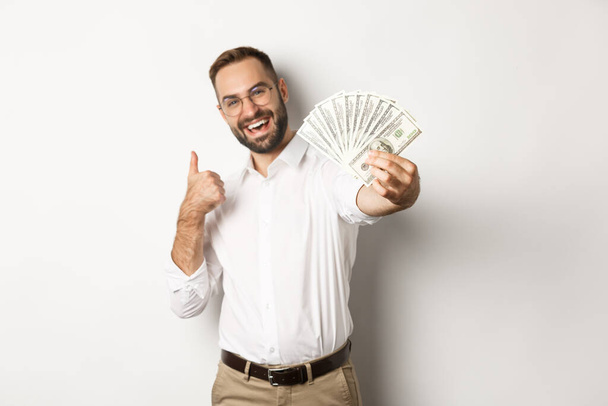 Επιτυχημένος επιχειρηματίας που δείχνει τα χρήματα δολάρια και τους αντίχειρες-up, χαμογελώντας ικανοποιημένοι, στέκεται πάνω από το λευκό φόντο - Φωτογραφία, εικόνα