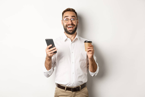 Amazed businessman drinking coffee, reacting at awesome online offer on mobile phone, standing over white background - Zdjęcie, obraz