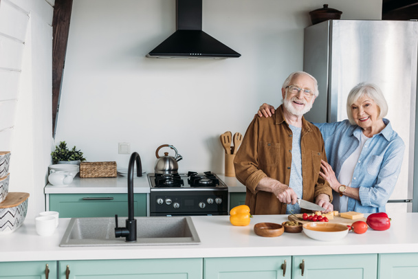 χαμογελαστή ηλικιωμένη σύζυγος κοιτάζει την κάμερα ενώ αγκαλιάζει τον σύζυγο μαγειρεύοντας δείπνο στην κουζίνα - Φωτογραφία, εικόνα