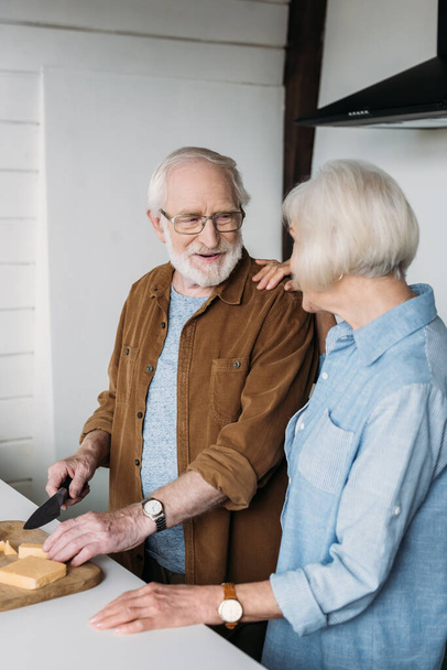 glimlachende senior man op zoek naar vrouw tijdens het snijden van kaas op snijplank in de keuken  - Foto, afbeelding