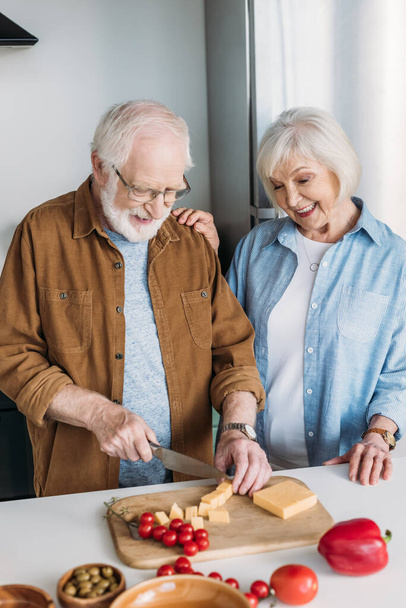 ευτυχισμένη ηλικιωμένη σύζυγος κοντά σύζυγος κοπής τυριού σε ξύλο κοπής κοντά σε λαχανικά στην κουζίνα - Φωτογραφία, εικόνα