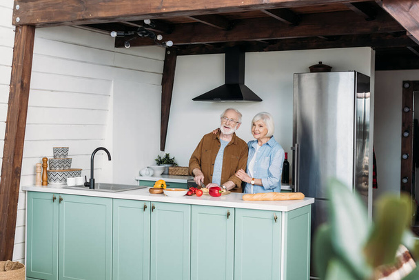 усміхнена старша жінка обіймає чоловіка, що ріже сир на столі з овочами та багет на кухні на розмитому фоні
 - Фото, зображення