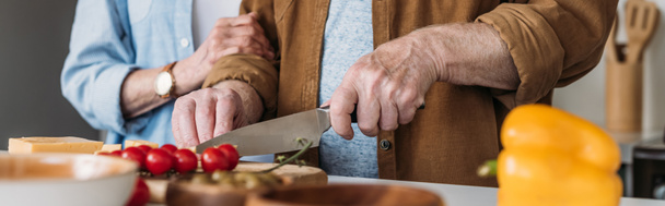 cropped view of elderly woman near man cutting cheese on table with vegetables on blurred foreground, banner - Photo, Image