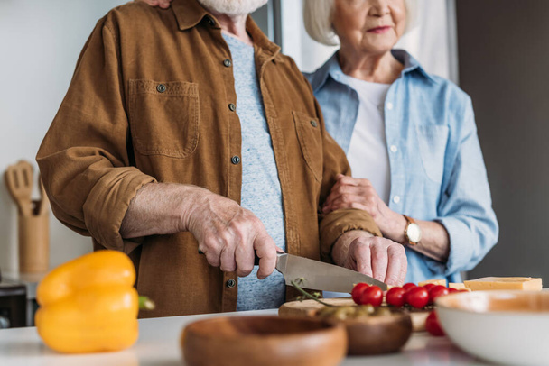 обрезанный вид пожилой жены рядом с мужем режет помидоры черри на кухне на размытом переднем плане - Фото, изображение