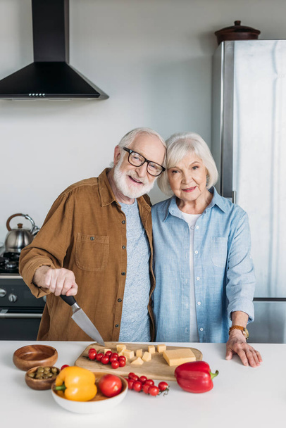 glückliches älteres Paar blickt in die Kamera, während es sich in der Küche am Tisch mit Essen umarmt - Foto, Bild
