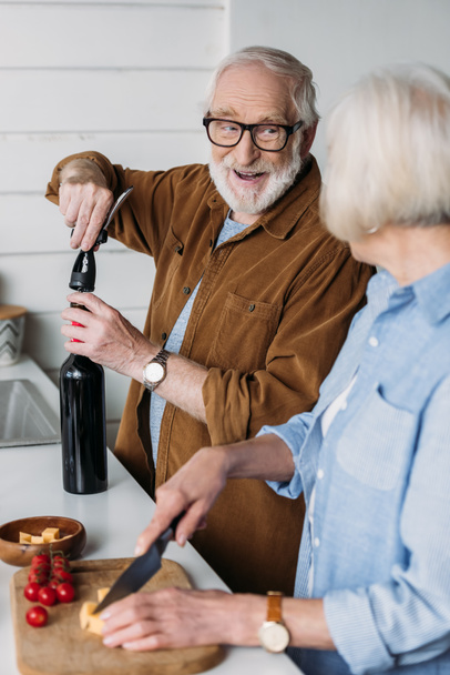 szczęśliwy starszy mąż patrząc na żonę podczas otwierania butelki wina korkociągiem w kuchni na zamazanym pierwszym planie - Zdjęcie, obraz