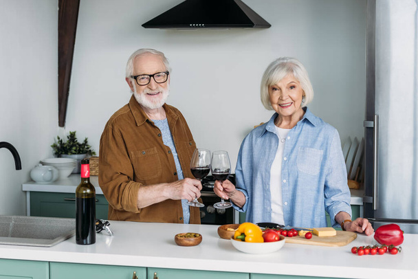 happy elderly couple looking at camera while toasting with wine glasses near table with food in kitchen - Photo, image