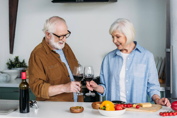 szczęśliwy senior para z kieliszkami do wina toasting w pobliżu stołu z jedzeniem w kuchni - Zdjęcie, obraz