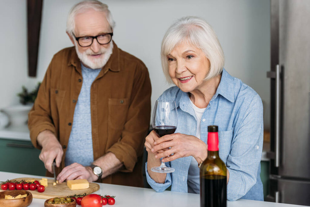 smiling senior husband looking at wife with wine glass while cutting cheese on chopping board in kitchen on blurred background - Photo, image