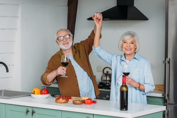szczęśliwy senior para z kieliszkami wina patrząc na aparat podczas zabawy w pobliżu stołu z jedzeniem w kuchni - Zdjęcie, obraz