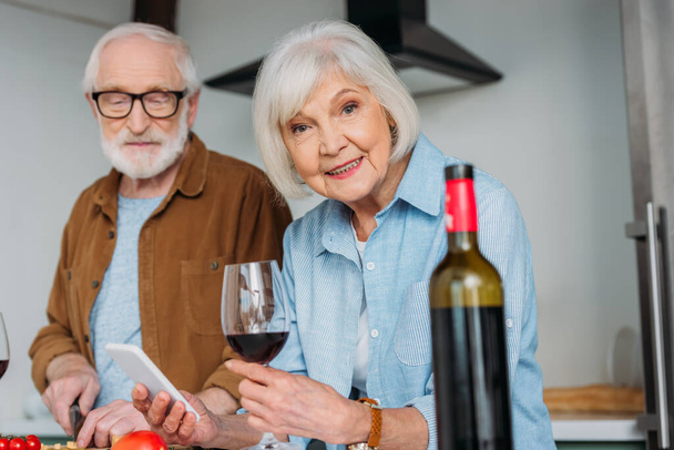 smiling senior woman with smartphone and wine glass looking at camera near husband with blurred bottle on foreground indoors - Photo, Image