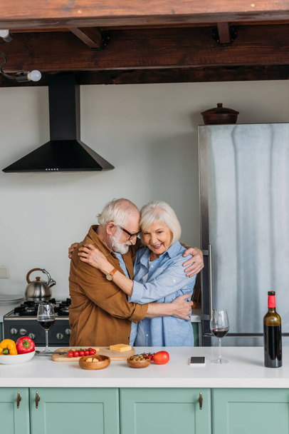 幸せな高齢者のカップルはキッチンで食べ物やワイングラスとテーブルの近くに抱きつく  - 写真・画像