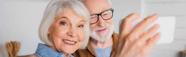 alegre mujer mayor mirando a la cámara mientras toma selfie con el hombre en primer plano borrosa en interiores, pancarta - Foto, Imagen