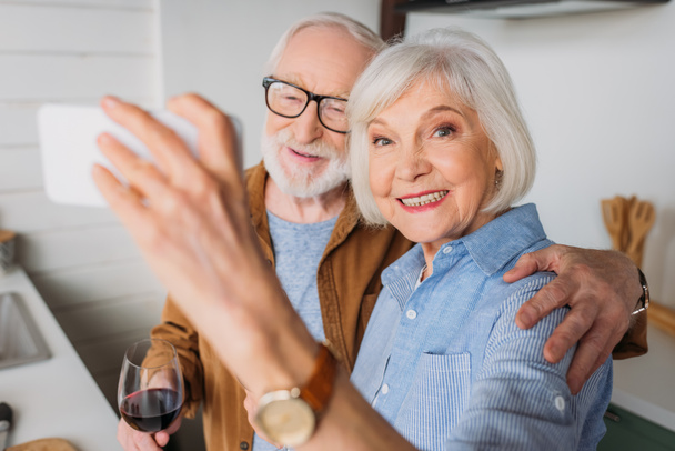 feliz anciana esposa mirando a la cámara mientras toma selfie con marido sosteniendo copa de vino en el interior en primer plano borrosa - Foto, imagen