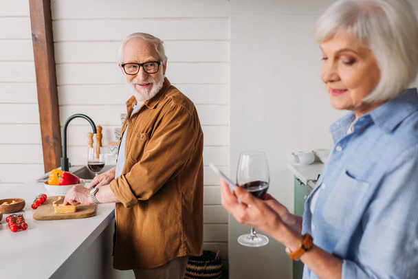 sonriente hombre mayor mirando a la mujer mientras se corta queso en la tabla de cortar en la cocina en primer plano borrosa - Foto, imagen