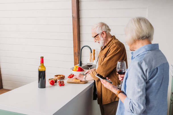 anciana esposa con teléfono inteligente y copa de vino cerca de marido sonriente cocinar la cena en la cocina en primer plano borrosa - Foto, imagen