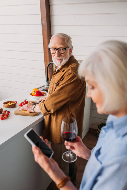 前景がぼやけてキッチンで夕食を調理しながら妻を見て笑っている高齢者の夫 - 写真・画像