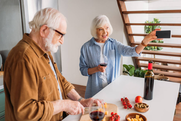 boldog idős feleség borospohárral selfie közel férj főzés vacsora a konyhában - Fotó, kép