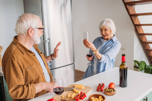 happy elderly wife with wine glass and smartphone taking photo of husband cooking dinner in kitchen - Photo, Image
