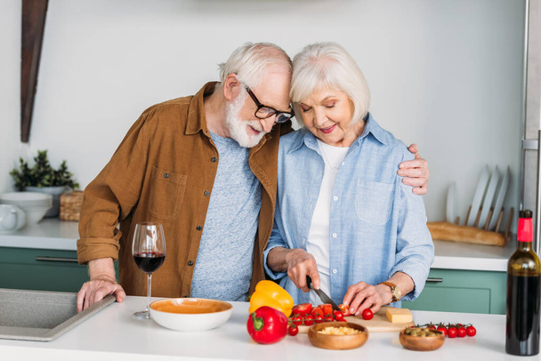 smiling senior husband hugging wife cooking dinner on table with wine, cheese and vegetables on blurred background - Photo, Image