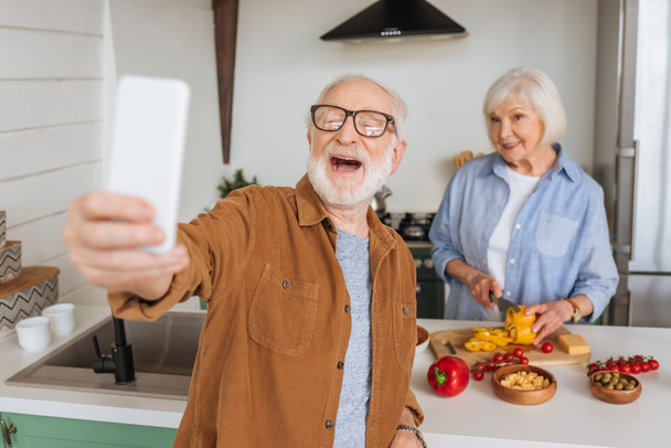 szczęśliwy starszy mąż biorąc selfie z żoną gotowanie obiad w kuchni na zamazanym pierwszym planie - Zdjęcie, obraz