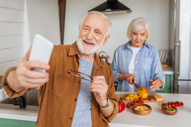 szczęśliwy starszy mąż ze smartfonem biorąc selfie z żoną gotowanie obiad w kuchni na zamazanym pierwszym planie - Zdjęcie, obraz