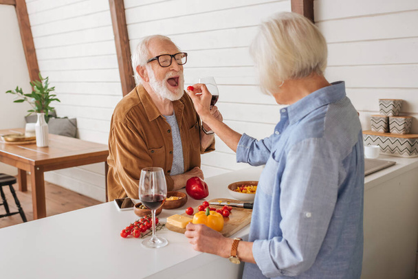 Rückansicht der älteren Frau, die ihren glücklichen Mann mit Kirschtomaten am Tisch mit Essen in der Küche füttert - Foto, Bild