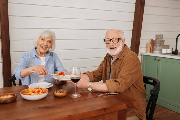 усміхнена старша дружина дивиться на камеру, подаючи салат чоловікові за столом з вегетаріанською вечерею на кухні
 - Фото, зображення