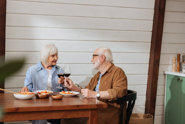 Lächelndes Seniorenpaar schaut sich beim Anstoßen mit Weingläsern am Tisch mit vegetarischem Abendessen in der Küche an - Foto, Bild