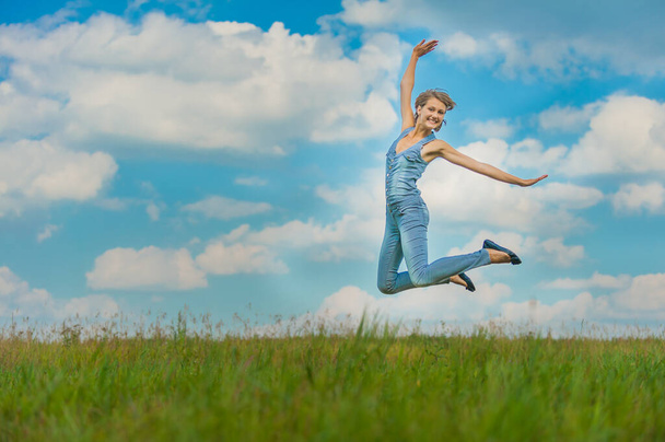 Jovem mulher bonita em um macacão de ganga azul diversão salta contra o céu azul com nuvens e grama verde ano de altura. - Foto, Imagem