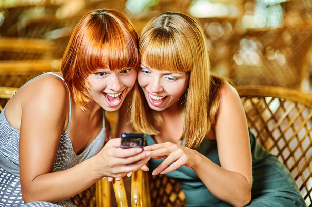 Dwie wesołe kobiety czytają smsy na telefonie komórkowym na tle dużej sali z wiklinowymi krzesłami. - Zdjęcie, obraz