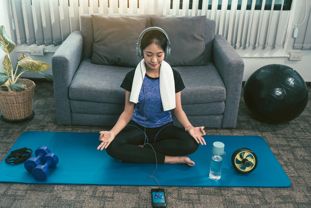 Azji piękna młoda kobieta za pomocą smartfona i słuchawek podczas medytacji i praktykowania jogi w domu. - Zdjęcie, obraz
