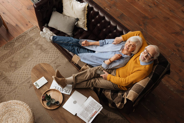 top view of senior couple looking at camera while hugging on couch in living room - Photo, Image