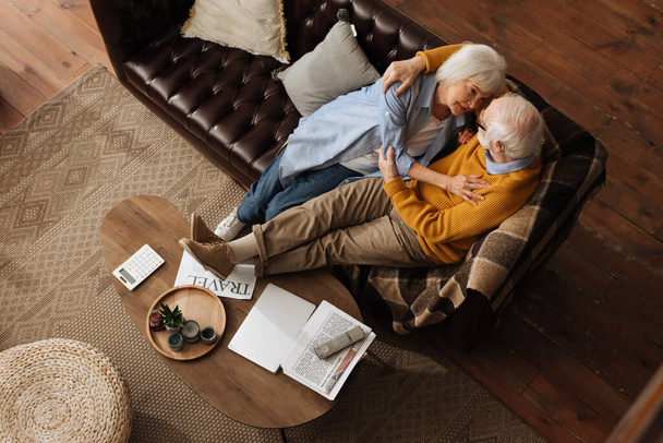 vista superior de la feliz pareja de ancianos mirándose mientras se abrazan en el sofá en casa - Foto, Imagen