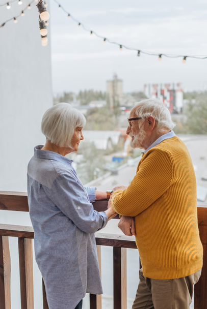 Lächelnder älterer Mann hält Hand in Hand mit Frau auf der Terrasse vor verschwommenem Hintergrund - Foto, Bild