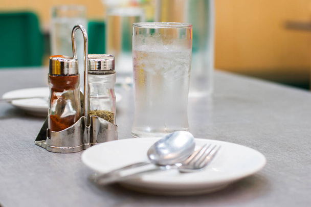 A cold glass of ice water complete, with plates, spoons, forks, and Sauce bottle, pepper bottle the dining table in a modern restaurant and light blurred in the restaurant background. - Photo, Image