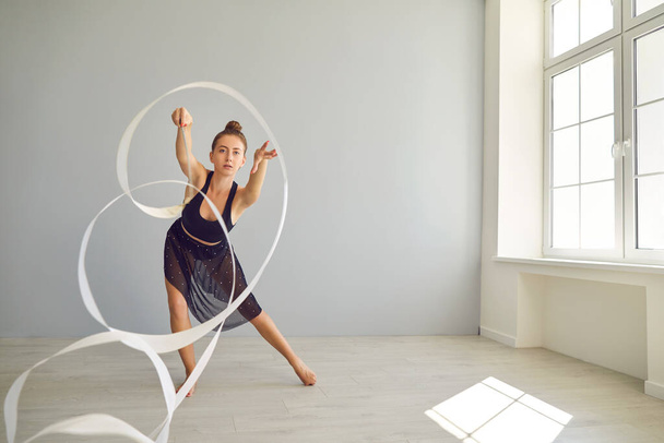Young rhythmic gymnast rehearsing movements with ribbon on warm floor in modern studio - Фото, зображення